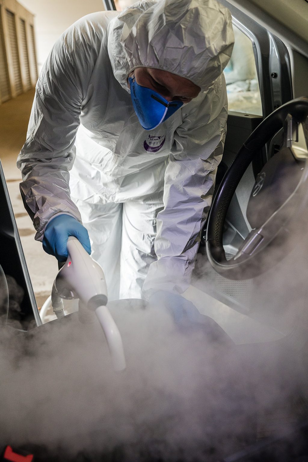 Persoon in beschermingspak en blauwe masker desinfecteert voertuiginterieur met sprayfles.