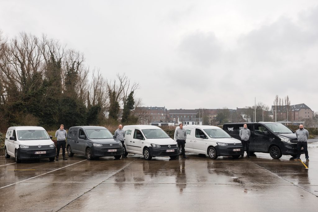 Een rij van zes Volkswagen busjes en bestelwagens staat opgesteld op een natte parkeerplaats, met werknemers erachter.