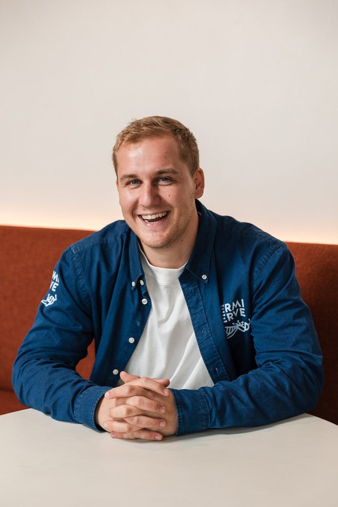 Een lachende man met rood-blond haar, gekleed in een blauw denim jasje over een wit shirt, zittend aan een tafel.